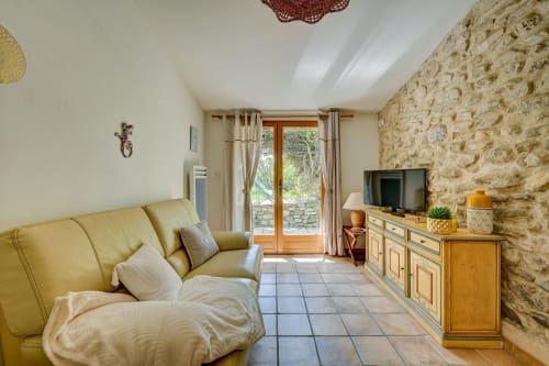 Séjour lumineux du gîte Agave avec canapé jaune, mur en pierre, téléviseur et portes ouvertes sur le jardin au Domaine Bellavigne