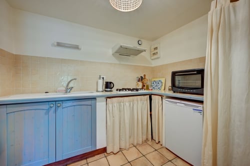 Cuisine aux armoires bleues avec carrelage beige, cuisinière, micro-ondes et rideaux crème au gîte Agave de Domaine Bellavigne