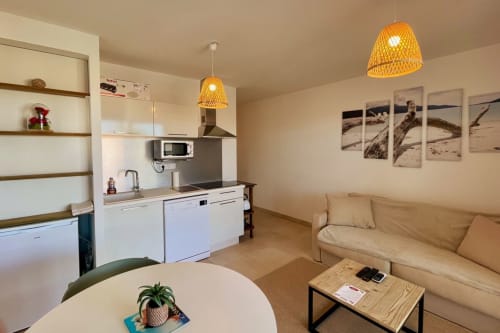 Accogliente monolocale T2 a Résidence Marina di Bravone con cucina a vista, divano beige, lampade sospese intrecciate e arredi costieri.
