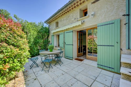 Terrasse ensoleillée du Domaine Bellavigne Gîtes avec bâtisse en pierre, volets verts, table à manger et jardins luxuriants à Forcalquier