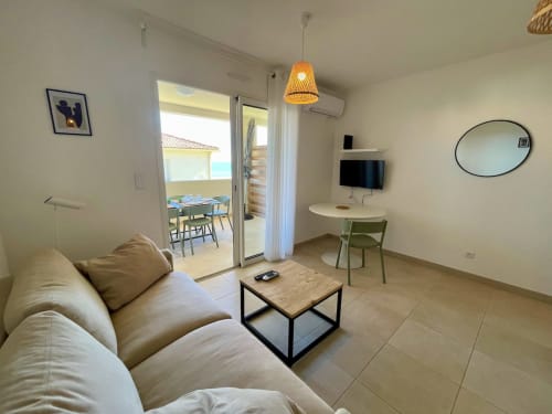 Elegante soggiorno con vista mare a Résidence Marina di Bravone, divano beige, zona pranzo, TV e accesso al balcone