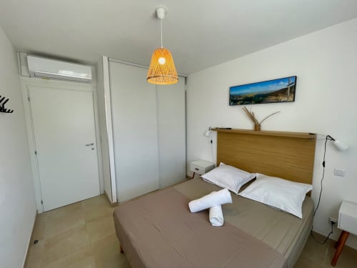 Chambre moderne à la Résidence Marina di Bravone avec lit en bois, lampe suspendue orange et vue côtière panoramique au-dessus de la tête de lit.