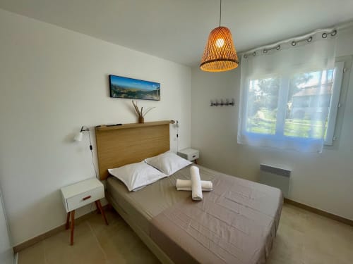 Chambre moderne à la Résidence Marina di Bravone avec lit en bois, luminaires suspendus et vue sur le jardin par portes vitrées.