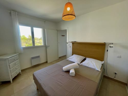 Camera luminosa a Résidence Marina di Bravone con letto in legno, cuscini bianchi, lampada a sospensione arancione e vista sul giardino