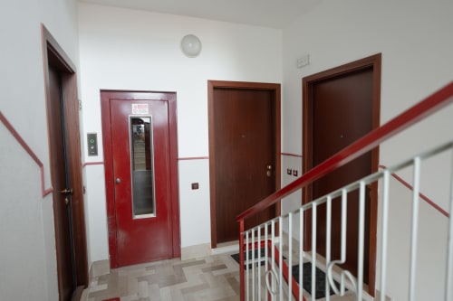 Corridoio della struttura con ascensore dalle porte rosse, porte di appartamenti in legno marrone e ringhiera bianca al Welcome Holiday di Finale