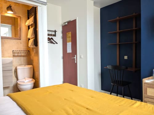 Chambre d'hôtel lumineuse avec lit jaune, mur d'accent bleu foncé, bureau en bois et salle de bains privative aux Maisons Delphine