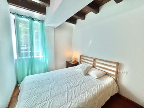 Dormitorio luminoso con cortinas turquesas, cabecero de listones de madera, ropa de cama blanca, vigas vistas y vistas a las montañas en Vielha