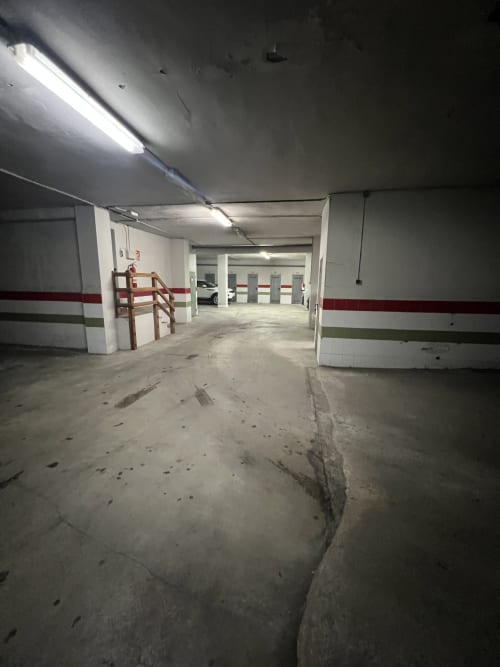 Garaje de estacionamiento subterráneo con paredes blancas, franja roja, iluminación fluorescente y vehículos estacionados en ERA CLAU HOMES