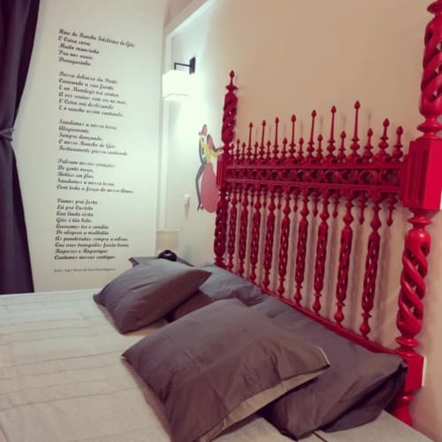 Quarto luminoso com cama de madeira vermelha ornamentada, almofadas cinzentas e poesia portuguesa nas paredes da Quinta SOTAM em Góis