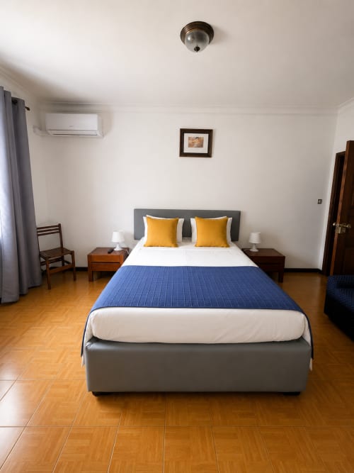 Quarto moderno na Serrana Gerês Inn com cama de casal, cabeceira cinza, almofadas amarelas, pisos de madeira e paredes brancas