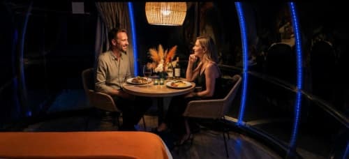 Couple dînant dans une salle bulle illuminée de l'hôtel Bubble Jade, avec vue sur jardin et arrangement central de pampas