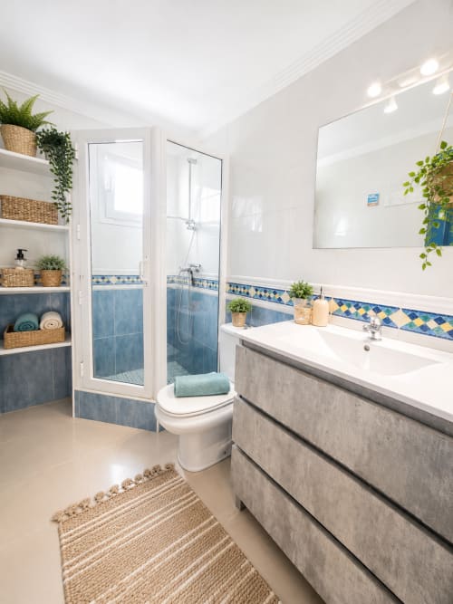 Modernes Badezimmer mit blauen Fliesenelementen, weißem Waschbeckenunterschrank, Glasduschwand und geflochtenen Körben in Corralejo-Apartment