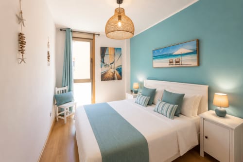 Habitación luminosa en Sara & Gino Fuerteventura con pared turquesa, ropa de cama blanca, lámpara colgante de mimbre y vistas al mar