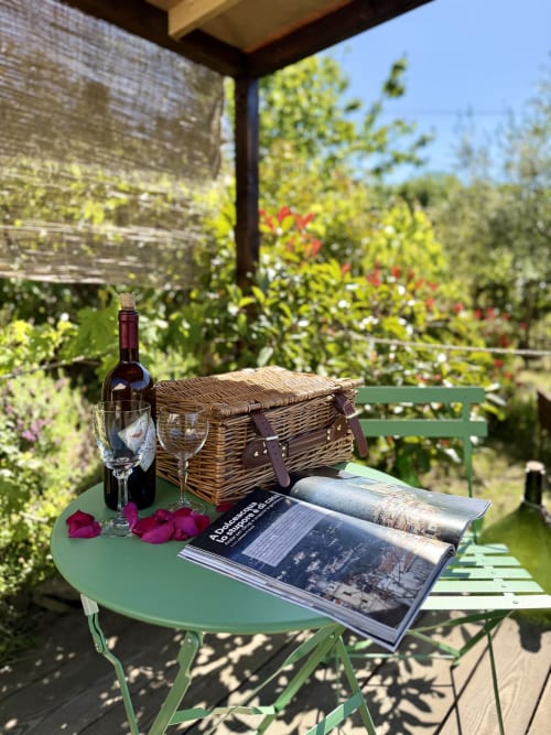 Mise en place pique-nique à l'Agritusmo Stecadò : bouteille de vin, verres, panier en osier et magazine sur table verte avec vue sur Dolceacqua