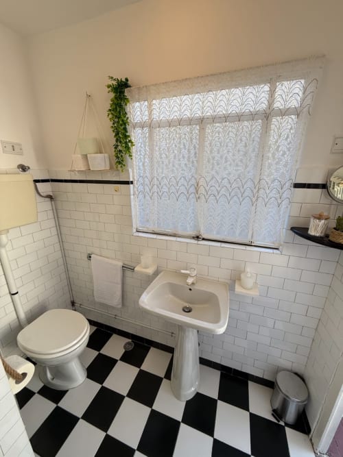 Bagno pulito con piastrelle bianche metro, pavimento a scacchiera bianco e nero, lavabo su colonna, toilette e tenda di pizzo alla finestra