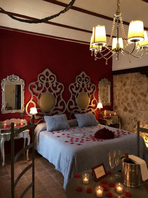 Romantic bedroom at Complejo Turístico Retiro del Teatro with red walls, ornate headboard, chandelier, and rose petals.
