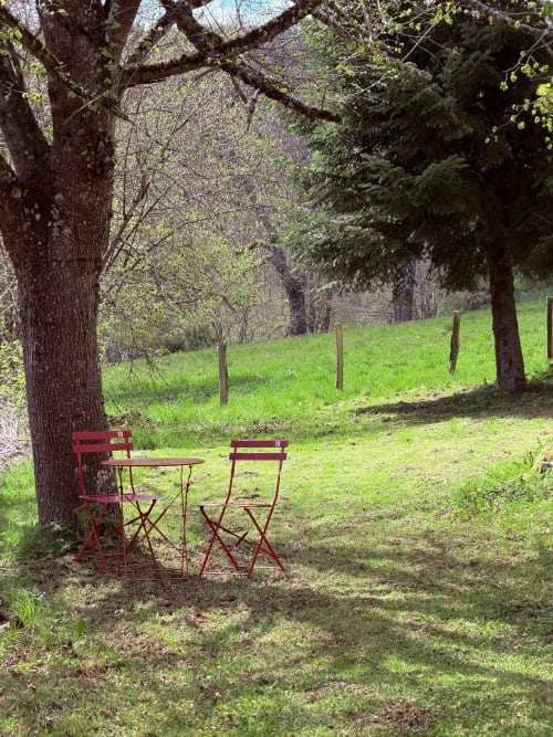 Chaises et table en métal rouge sous un grand arbre dans le jardin verdoyant de la villa Le 1830 à Églisolles