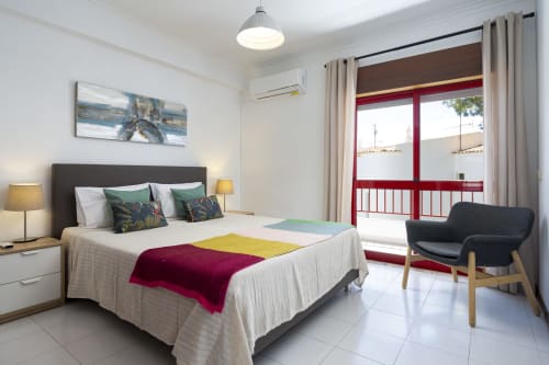 Quarto moderno em RB Algarve Vilamoura com cama de casal, manta colorida, porta-varanda vermelha, cadeira preta e luminária de teto