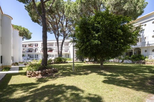 Pátio do hotel RB Algarve com árvores centenárias, jardim impecável e edifícios brancos em Vilamoura, Portugal