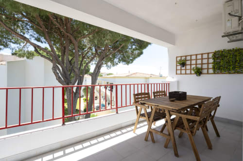 Varanda ensolarada do apartamento RB Algarve Vilamoura com mesa de refeições, guarda-corpo vermelho e grande árvore com vista para a piscina