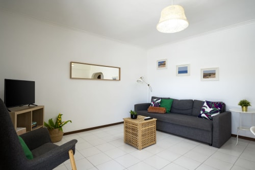 Sala de estar moderna no apartamento DINIS em RB Algarve Vilamoura com sofá cinzento, TV, móveis em madeira e piso em azulejos brancos