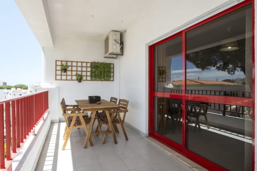 Varanda ensolarada do apartamento RB Algarve DINIS com mesa de jantar em madeira, portas vermelhas e vista para o mar em Vilamoura, Portugal