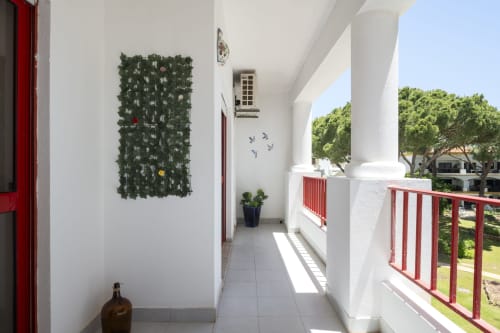 Varanda coberta do hotel RB Algarve com paredes brancas, grades vermelhas, jardim vertical verde, plantas em vasos e vista para a piscina