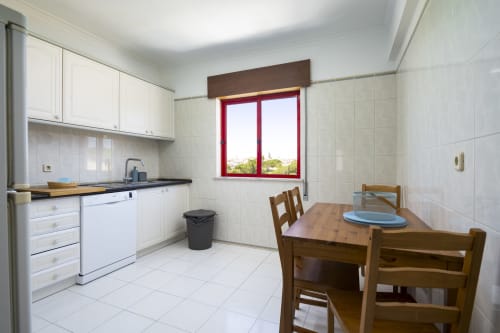 Cozinha e área de refeições do apartamento DINIS no RB Algarve, Vilamoura com armários brancos, janela com moldura vermelha e mesa de madeira