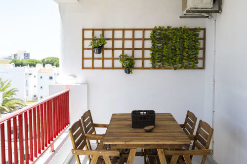 Área de refeições na varanda do apartamento DINIS RB Algarve, Vilamoura, com mesa de madeira, cadeiras, guarda-corpo vermelho e plantas suspensas.