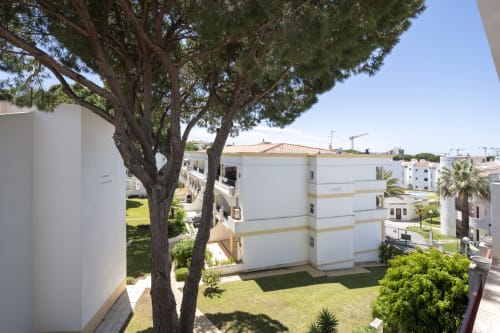 Exterior do apartamento RB Algarve em Vilamoura com edifícios brancos, pinheiros frondosos e jardins bem cuidados sob céu azul claro