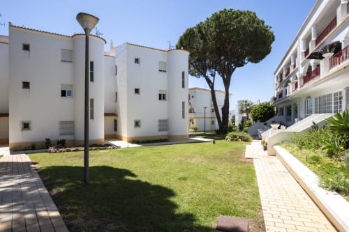 Pátio do complexo de apartamentos RB Algarve Vilamoura com edifícios brancos, gramado verde e árvore centenária