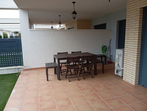 Patio couvert avec carrelage en terre cuite, table à manger en bois foncé, chaises, mur en briques blanches et façade en briques
