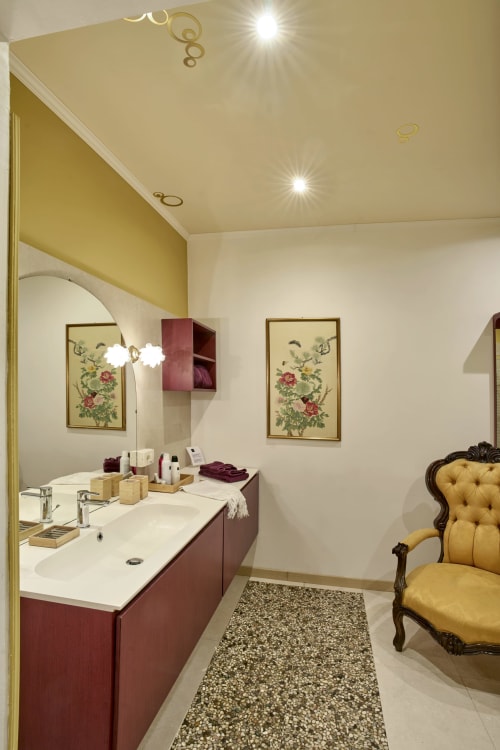 Elegante bagno con vanità bordeaux, top in marmo crema, opera botanica con cornice dorata e sedia rivestita in giallo