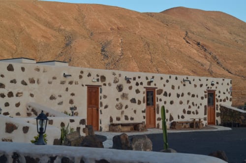 Hotel Rural El Cabo en Villaverde: edificio de piedra blanca con puertas rojas y fachada de roca volcánica sobre fondo marrón