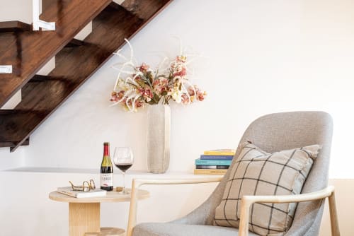 Elegante sala de estar do RB Algarve com poltrona bege, prateleiras de madeira, taça de vinho, flores secas em vaso e livros empilhados