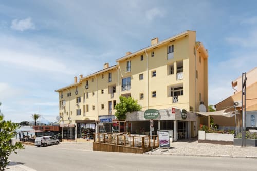 Edifício do hotel RB Algarve em Vilamoura com fachada amarela, lojas no piso térreo e localização à beira-mar sob céu azul