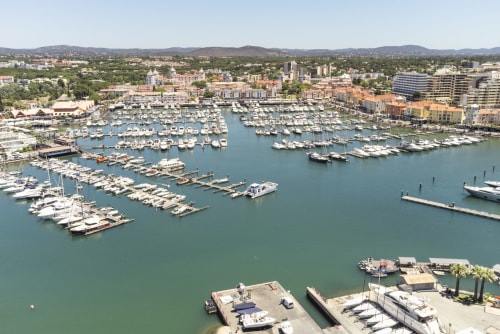 Vista aérea da marina de Vilamoura com centenas de barcos brancos atracados, edifícios coloridos à beira-mar e colinas verdes