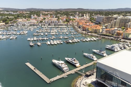 Marina com iates amarrados e barcos em Vilamoura, Portugal, com edifícios coloridos à beira-mar e colinas
