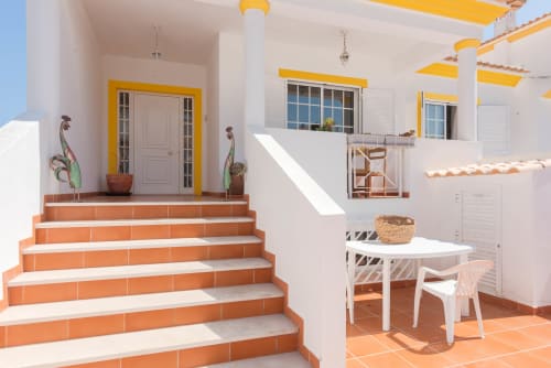 Terraço ensolarado da villa JUDY em RB Algarve com degraus laranja, acentos amarelos, mobiliário branco e plantas em vaso