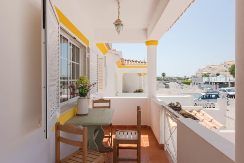 Terraço ensolarado da villa JUDY, RB Algarve, com cadeiras de madeira, mesa de jantar, detalhes em amarelo, flores em vasos e vista da piscina