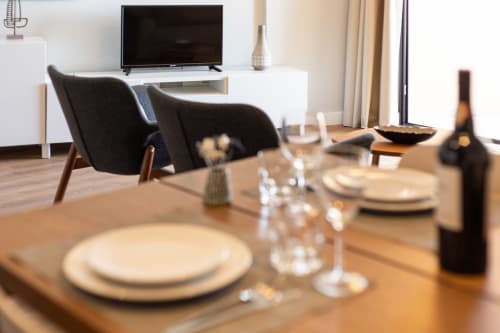 Área de jantar moderna no apartamento RB Algarve MILA com mesa posta, copos, garrafa de vinho e cadeiras pretas com vista para a sala
