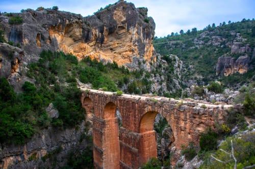 Puente acueducto romano antiguo atravesando cañón rocoso con exuberante vegetación verde bajo cielo azul