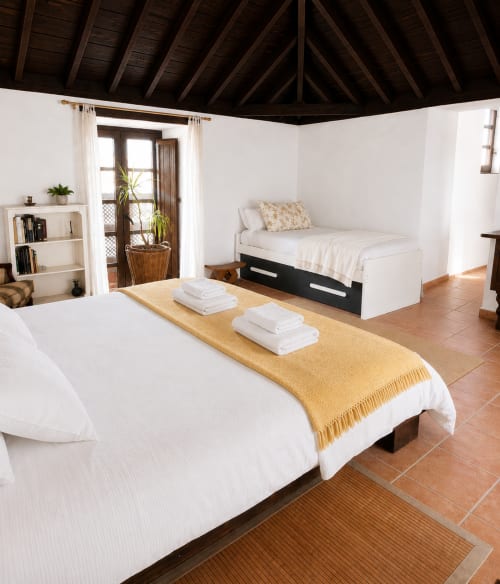 Chambre spacieuse à Sara & Gino Fuerteventura avec deux lits, draps blancs, plaid jaune, poutres en bois et vue jardin