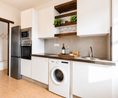 Cuisine moderne avec armoires blanches, poutres en bois, électroménagers inoxydables, lave-linge et verres à vin