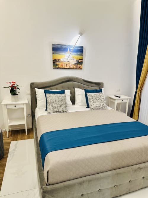 Letto matrimoniale deluxe con testata in tessuto grigio, accenti blu, comodini bianchi e vista città a St Mary Major Loft Roma