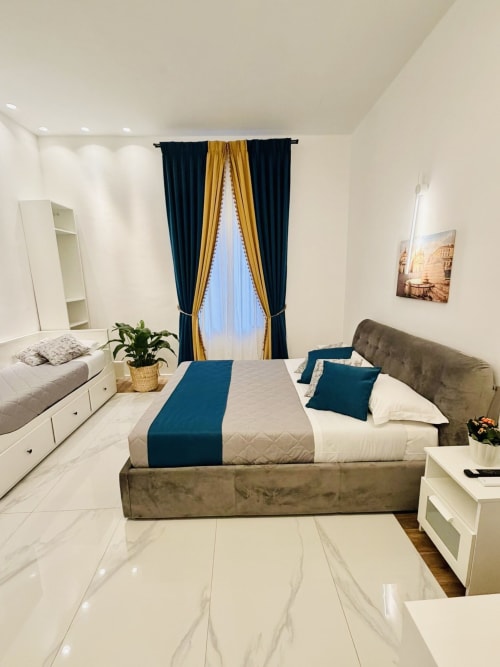 Elegante camera tripla al St Mary Major Loft Roma con letto imbottito grigio, accenti blu, tende raffinate e marmo
