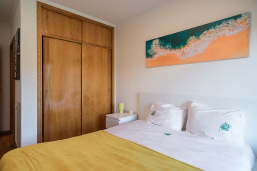 Quarto luminoso na EcoHost Gaia Beach&Golf com manta amarela, almofadas brancas, guarda-roupa em madeira e painel artístico de ondas costeiras