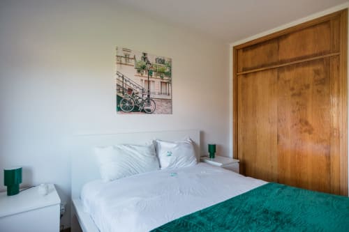 Quarto moderno com roupa de cama branca, manta verde, guarda-roupa em madeira e arte de bicicleta no apartamento EcoHost em Portugal