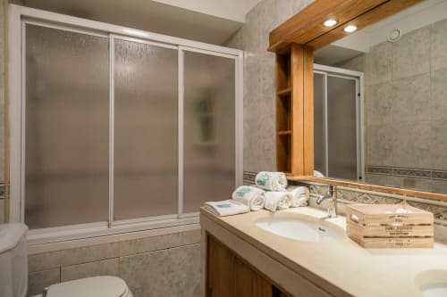 Banheiro moderno com pia dupla, acabamentos em madeira, box com vidro fosco, toalhas enroladas e detalhes decorativos em madeira