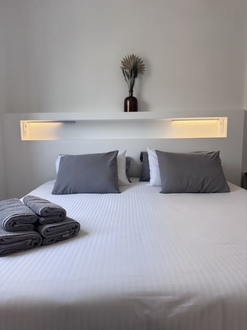 Dormitorio moderno en Kamalahouse, Corralejo, con cama blanca, almohadas oscuras, estante iluminado y toallas dobladas